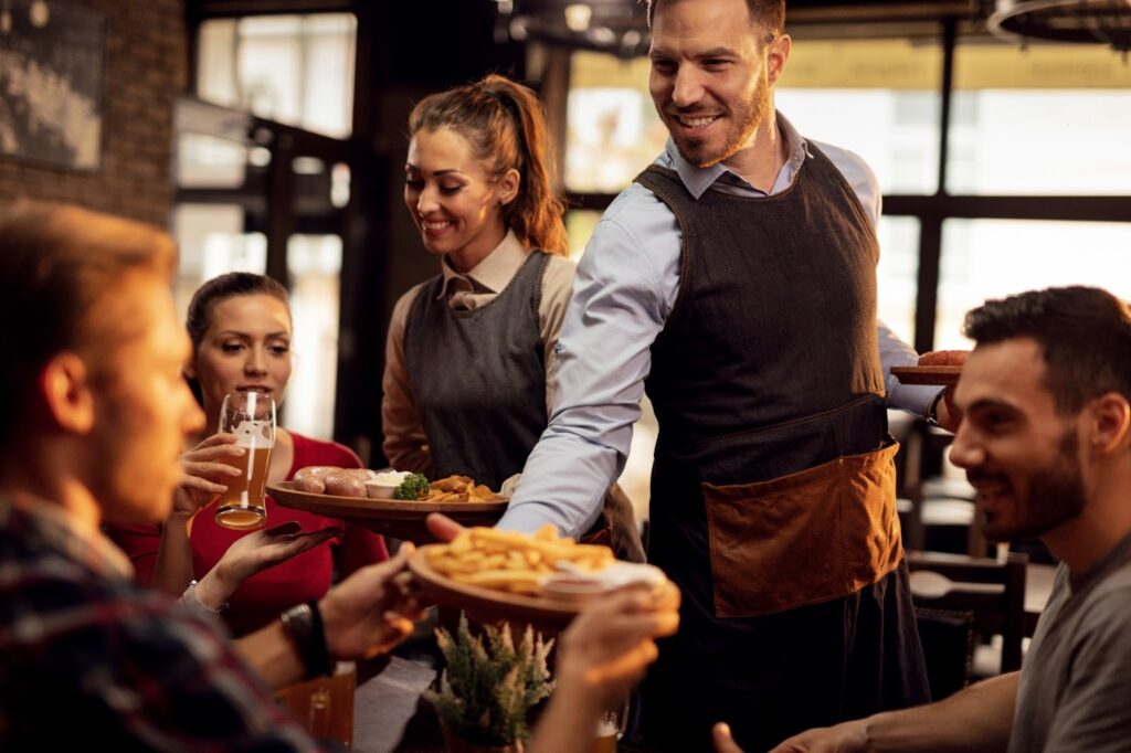 Happy waiters bringing food at the table and serving group of friends in a restaurant